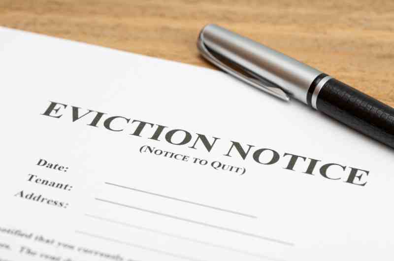 An older man opening the front door, staring into the camera as his landlord hands him an eviction notice.