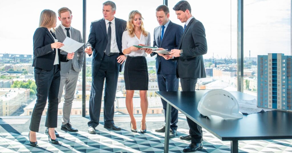 A group of six, well-dressed professionals looking over various documents as they stand in front of the window in their high-rise building.
