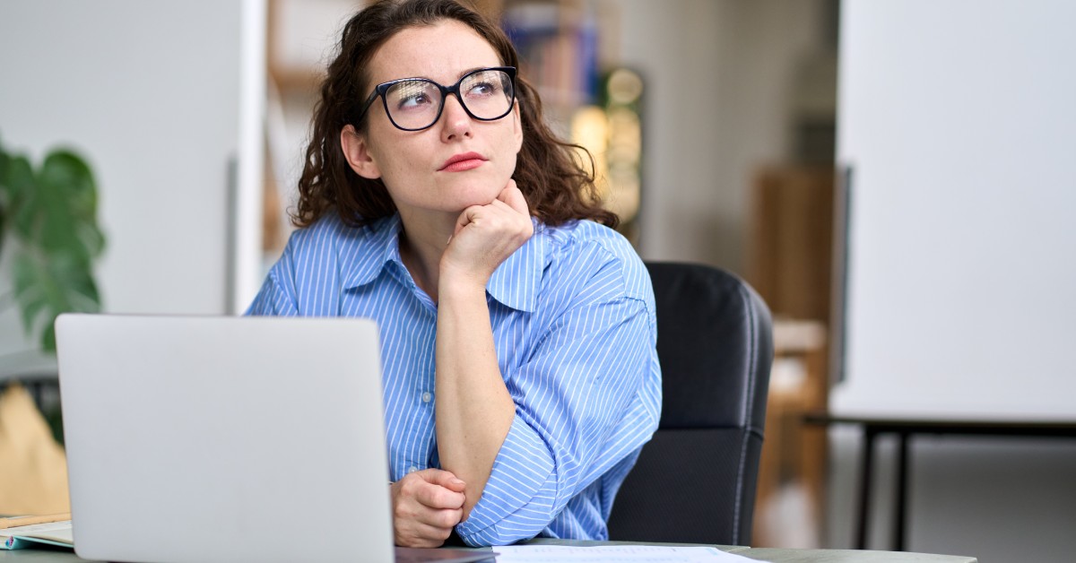 A businesswoman wearing glasses, sitting at her computer desk as she stares off thoughtfully into the distance.
