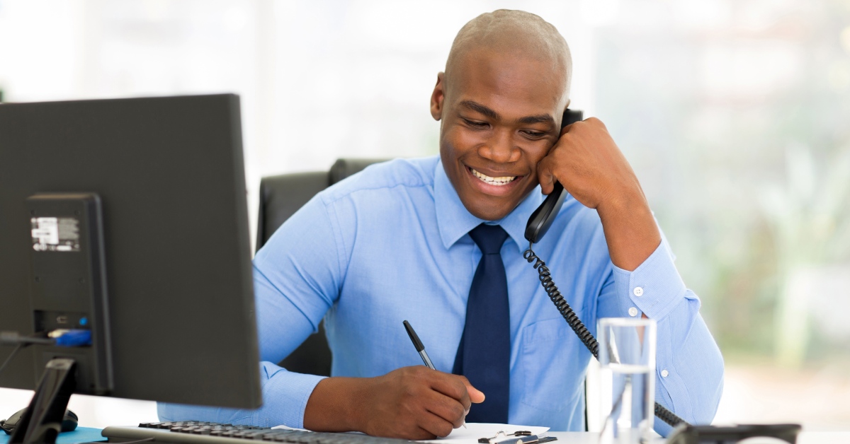 A businessman in a blue dress shirt and tie sitting at his computer desk while smiling and talking on the phone.