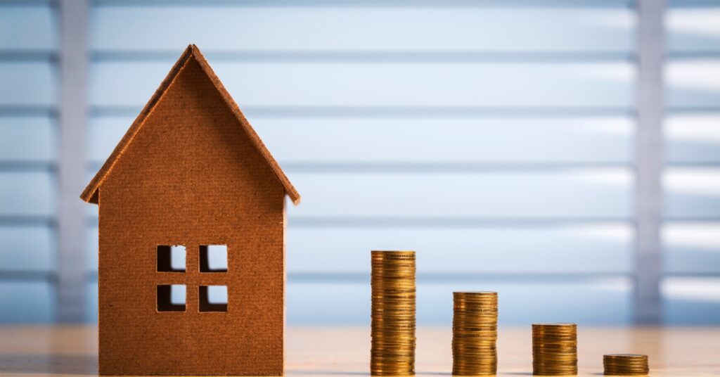 A tiny cardboard house model placed on a wooden desk next to an incrementally rising stack of coins.