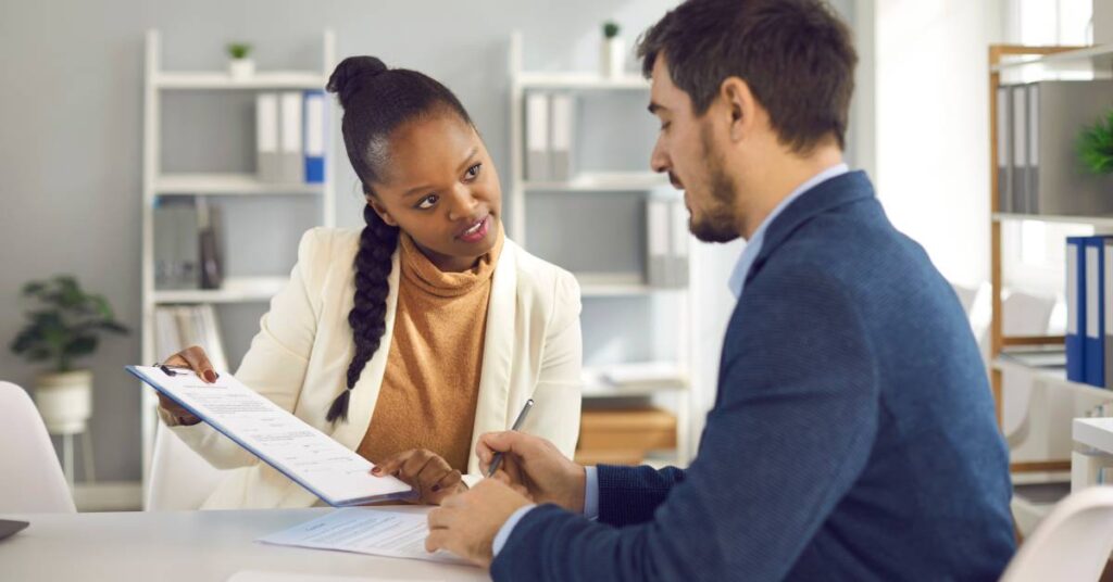 A female professional pointing and showing their male client a set of documents clipped to a blue clipboard.