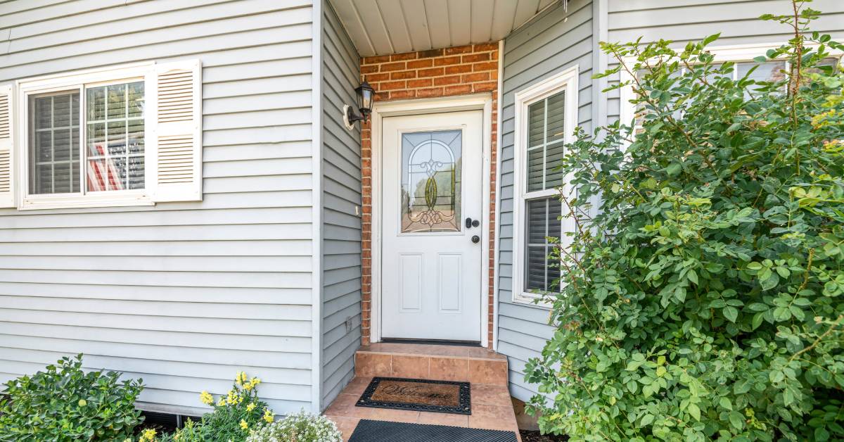 The front door of a home is surrounded by bushes with a welcome mat at the front step and a security camera above the doorknob.