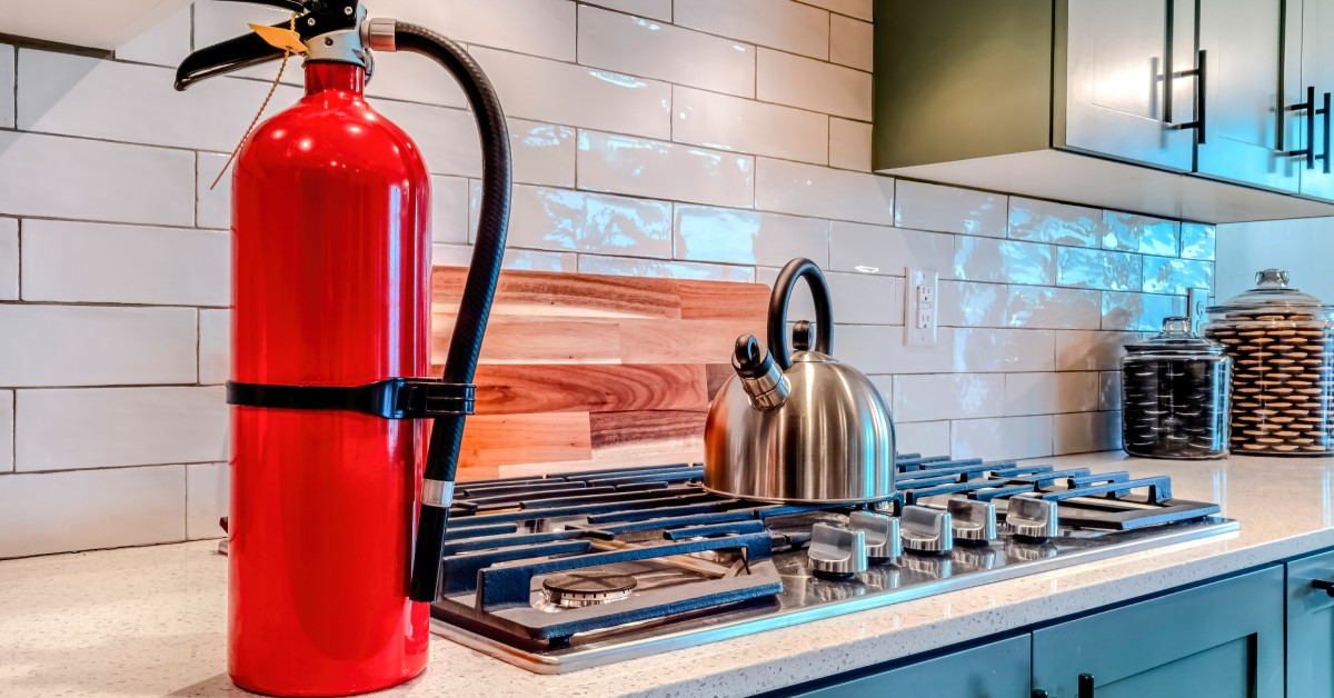 A classic, bright red fire extinguisher sitting on top of a kitchen counter next to a downdraft stove with a kettle on top.