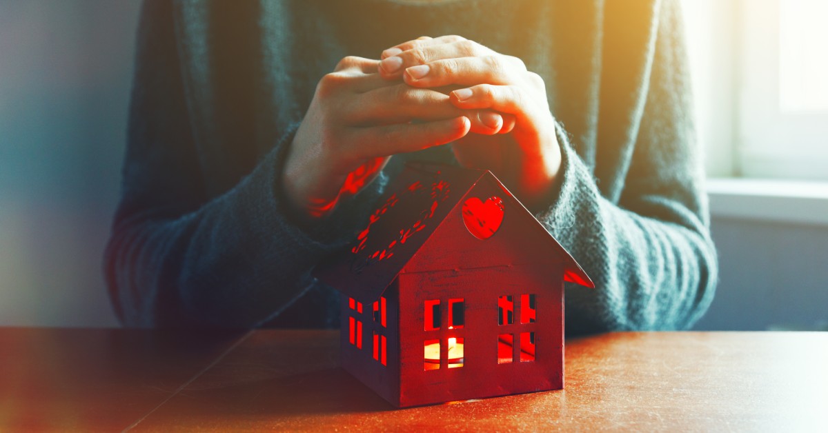 A pair of hands covering a small wooden model home with a heart carved into it as it glows red from the inside.