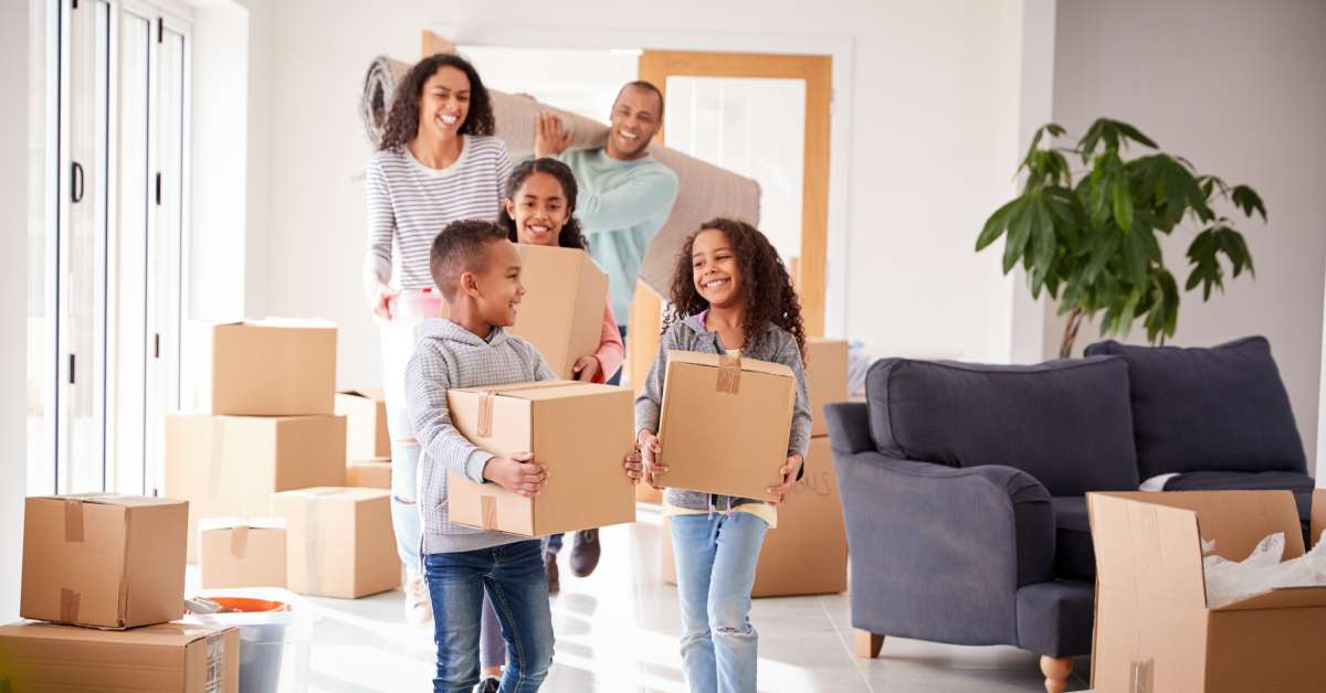 A smiling, happy family walking into their new home while the children and mother hold boxes and the father carries their rug.