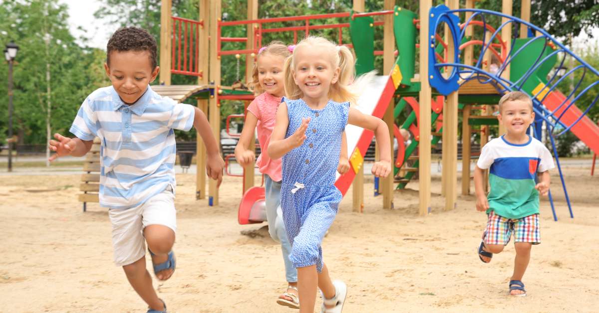 Happy, smiling children on a sandy play area running away from the colorful playground and slide set.