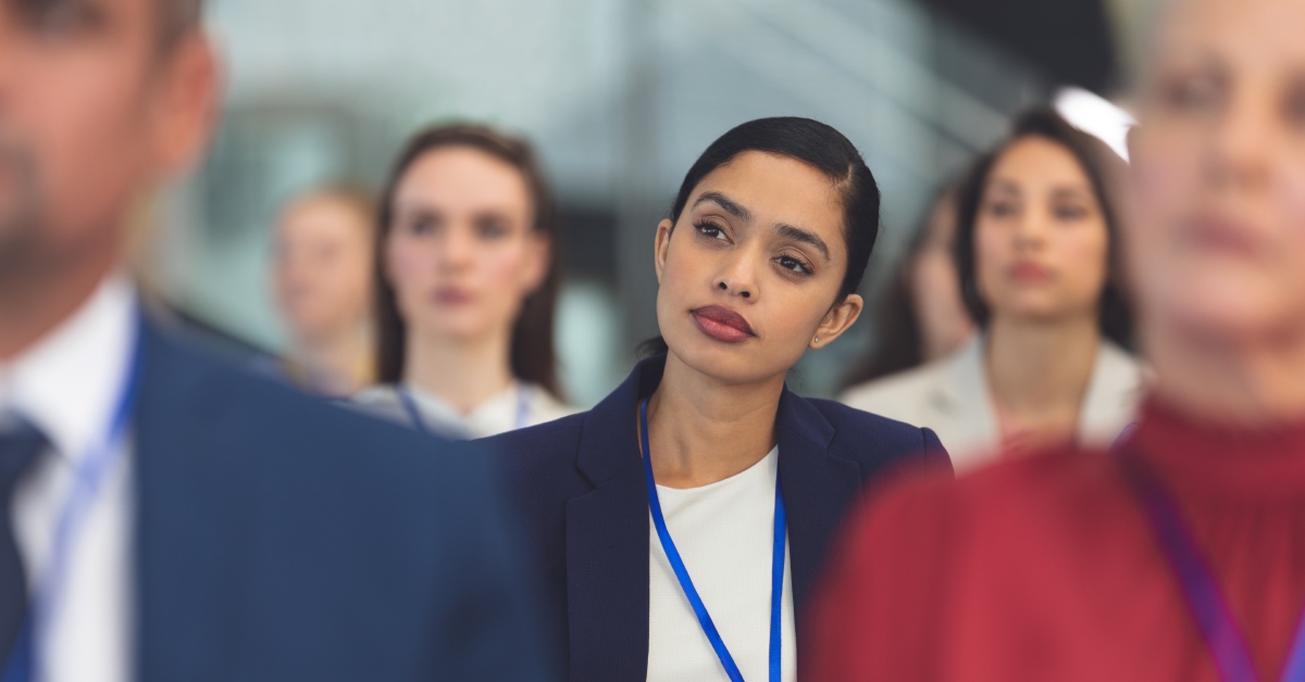 A businesswoman in a blue suit and lanyard sitting in the middle of a seminar, looking forward with a thoughtful expression.