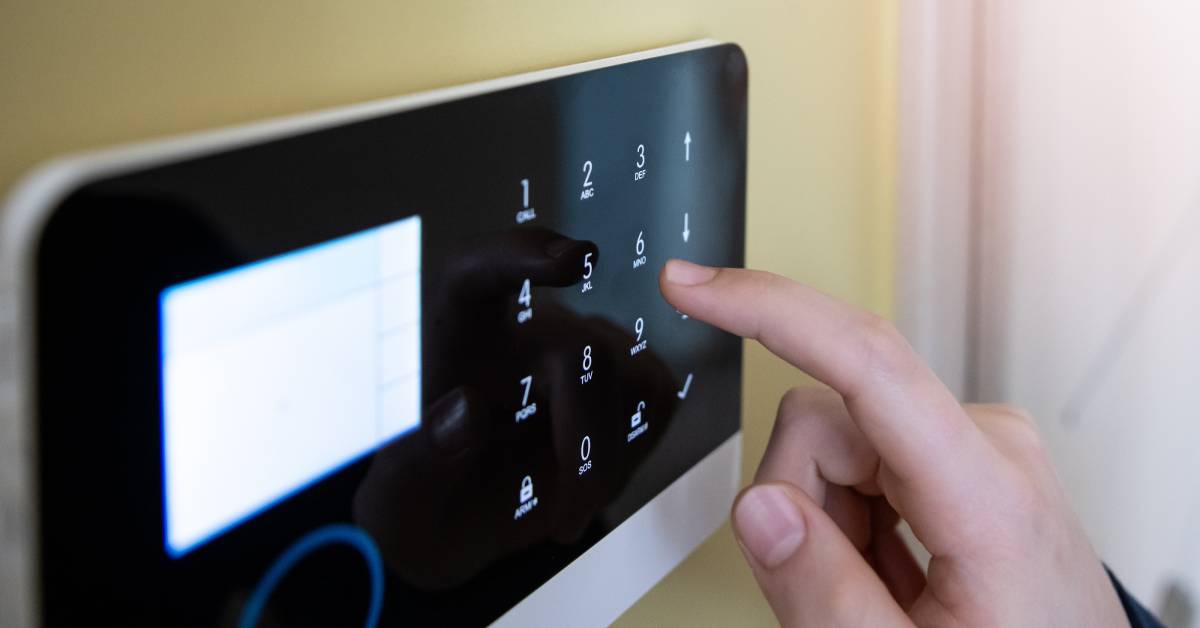 A man's hand reaching up to type in the security code on a black and white alarm system hanging on a wall.
