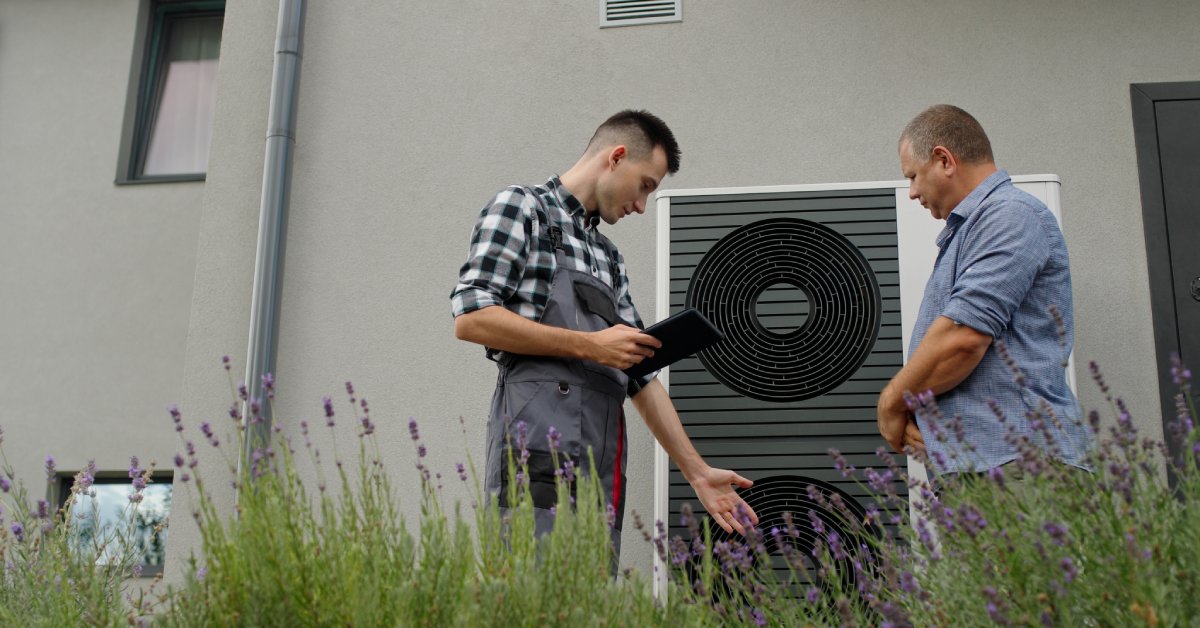 A technician in a grey Aaron and black and white flannel standing next to an AC unit, speaking to the man across from him.