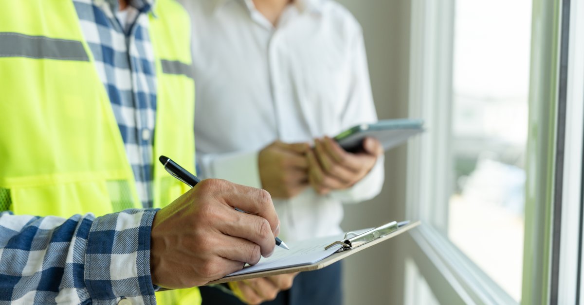 A close-up of an inspector in neon green high visibility gear standing next to a man in a white blouse who is holding a tablet.