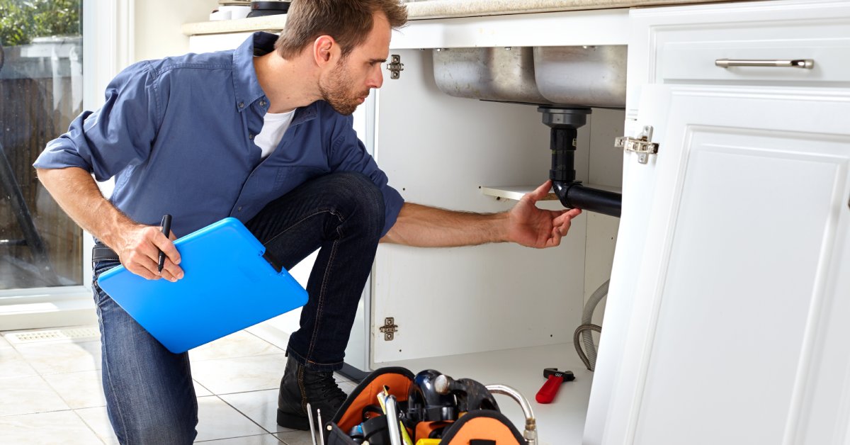 A man in jeans, a blue button-down shirt, and a white undershirt kneeling down to inspect the pipes under a sink.