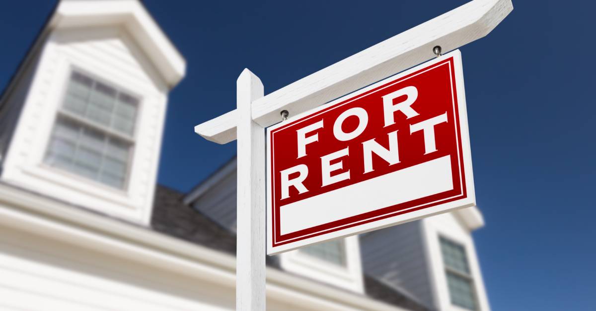 A red and white "For Rent" sign hanging on a white post in front of a two-story home, and blue sky in the blurred background.
