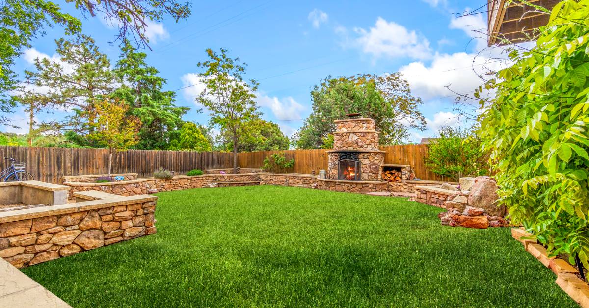 A wide-angle view of a well-landscaped backyard with a wood fence, stone retaining wall, and matching fireplace.