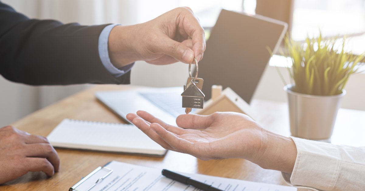 A man in a suit hands a house key to a person holding out their hand above a desk with a contract on a clipboard.