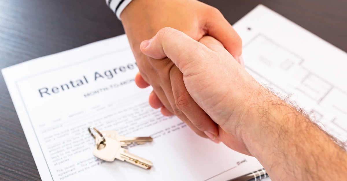 A close-up of two hands shaking over a contract reading "rental agreement". A pair of keys and a pen sit on top of the contract.