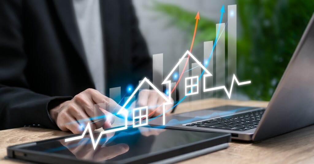A man sits at his laptop next to a tablet. An upward graph with homes representing real estate growth floats above the tablet.