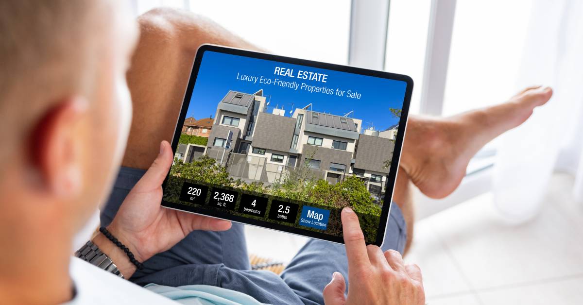 An over-the-shoulder view of a man viewing the property listing of a modern-two story home on a tablet.