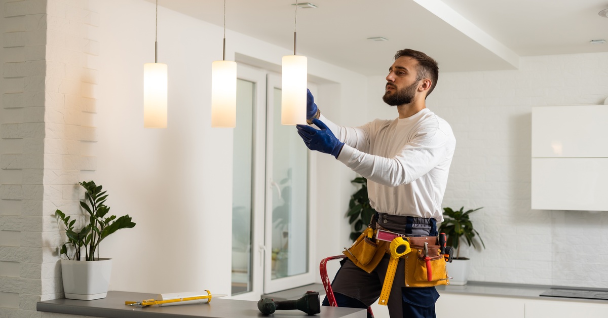 An electrician, wearing a tool belt and standing on a step ladder, installs a cylindrical pendant lamp over a kitchen counter.