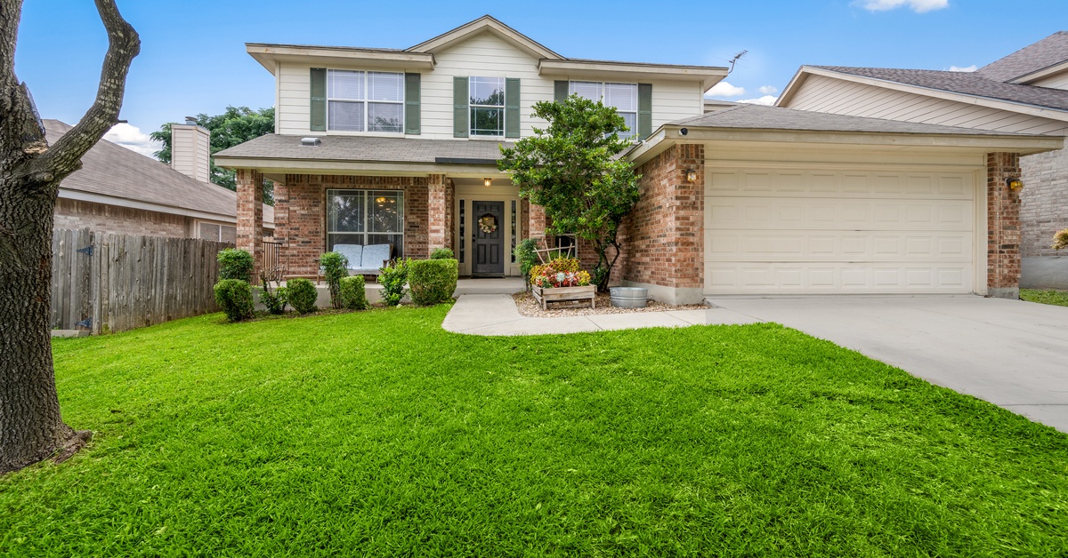 A two-story suburban home with a garage and an empty driveway. The bushes, front tree, and lawn are well-kept.