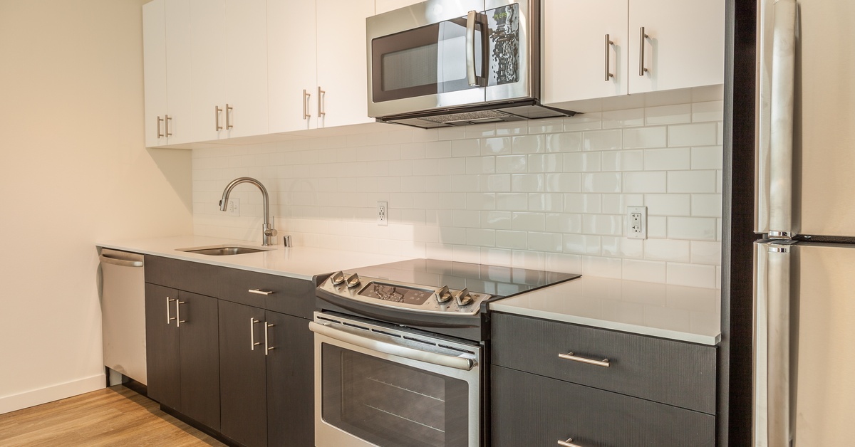 An apartment kitchen with stainless steel appliances and fixtures. There are white cabinets on top and gray cabinets below.