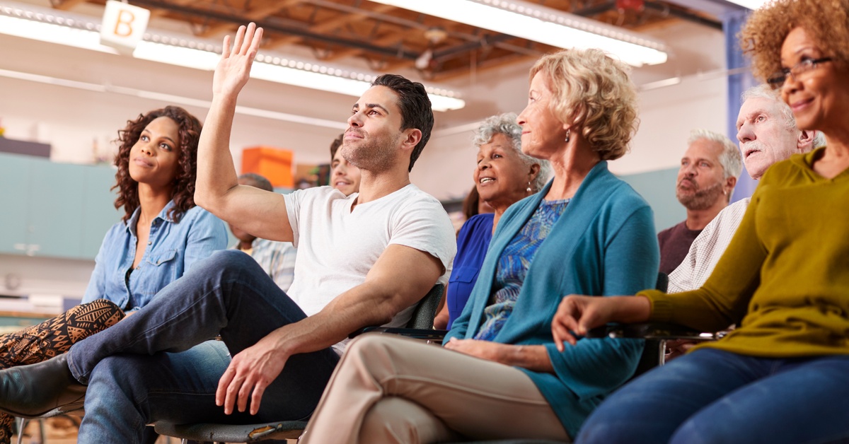 Members of a community meeting sit in rows of chairs. A man wearing a white t-shirt in the front row raises his hand.