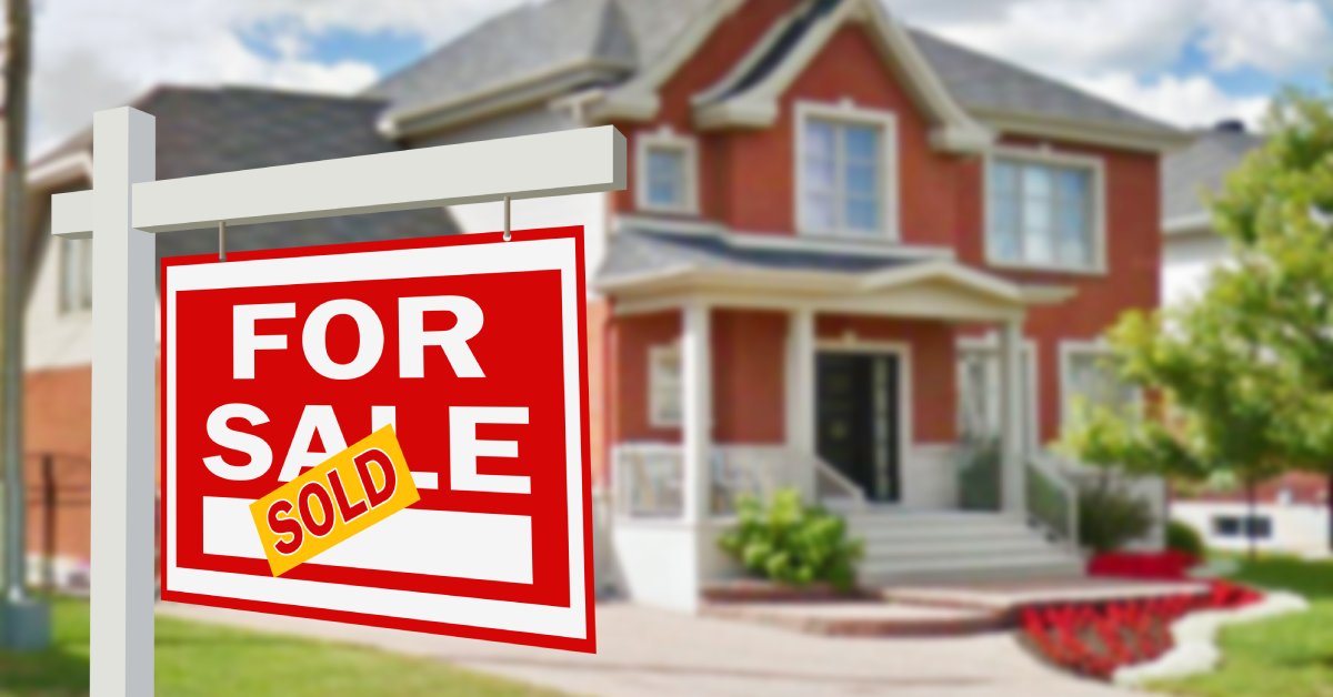 A red and white "for sale" sign covered with a yellow "sold" sticker hangs on a post in front of a two-story brick home.