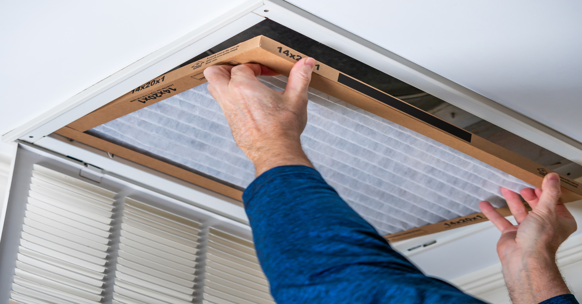 A man wearing a blue long-sleeved shirt installs a new HVAC air filter into a ceiling vent, with the vent panel hanging open.
