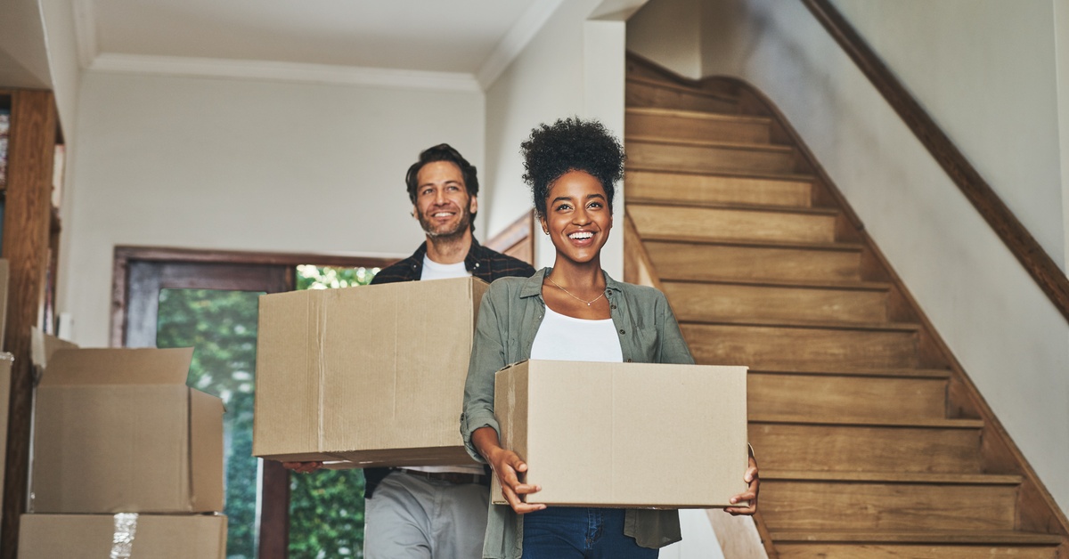 A male and a female both hold cardboard boxes as they walk down the hallway of a house. Boxes are stacked on one side.