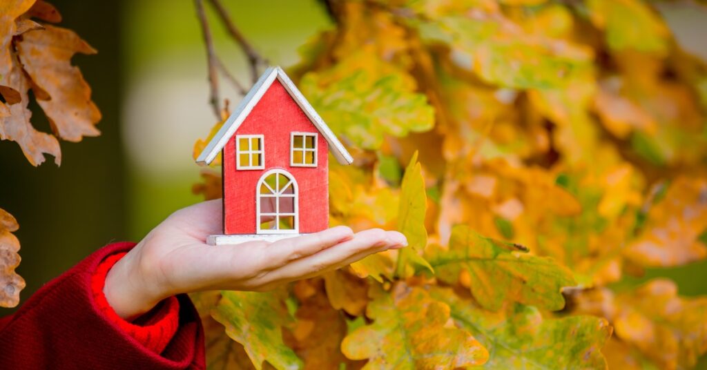 A hand holds a figurine of a red house with a white roof. Yellow and orange leaves of a tree fill the background.