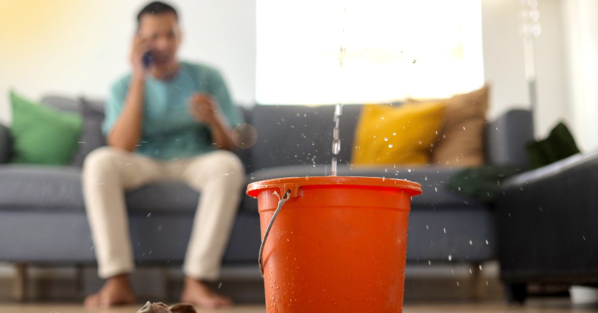 A man sits on a couch with a phone to his ear. In the foreground, there's a bucket catching water from a leak.