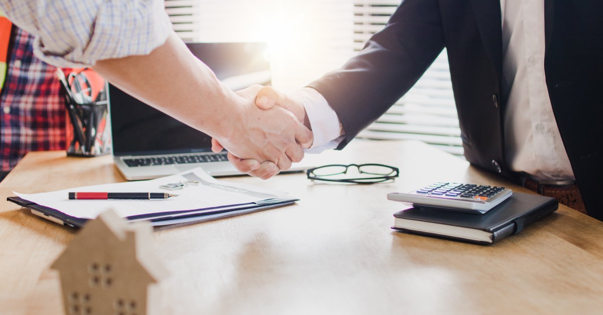 Above a table with a laptop and a wooden house model, two people shake hands. One person wears a suit.