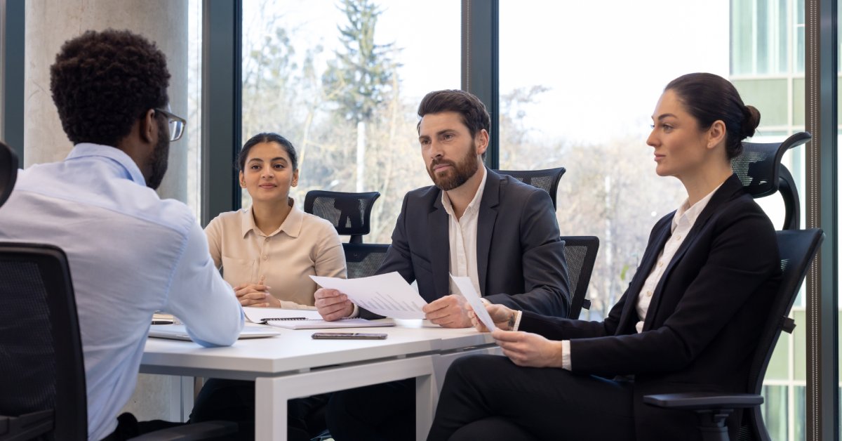 Four business professionals wearing business attire are sitting at a table and holding papers. The office has tall windows.