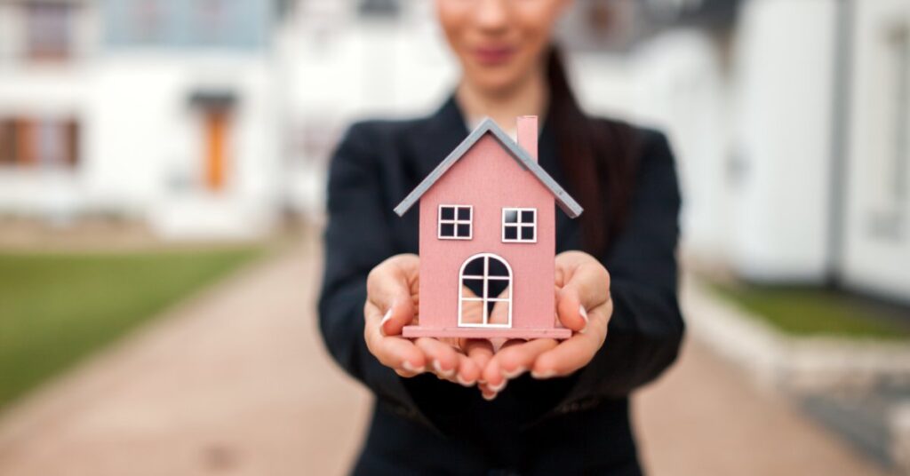 A woman wearing a black blazer holds a model of a red house. She stands in front of an apartment complex of white buildings.