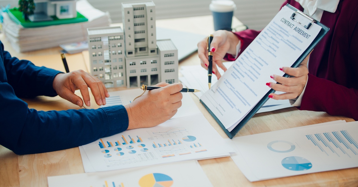 Two people sit at a table. One holds a contract agreement on a clipboard, and the other points at the paper with a pen.