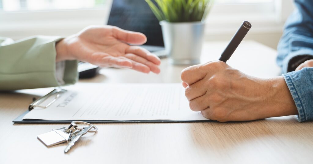 A person signs a paper attached to a clipboard beside a key. An individual sitting across the table holds out their hand.