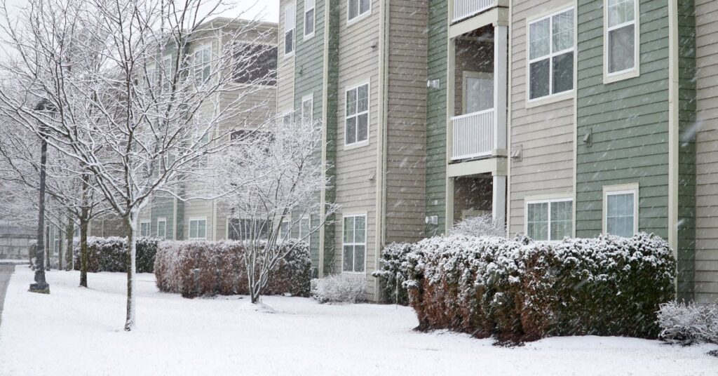 The exterior of an apartment building with three stories. Snow covers the ground and dusts the bare branches of a tree.