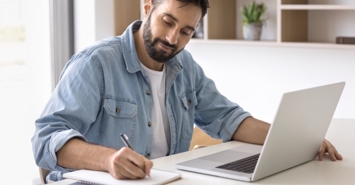 A man sits at a white desk that has two notebooks and a laptop on the surface. He writes on a blank sheet of paper.