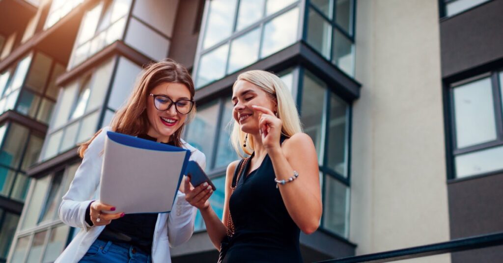 One woman holds a folder, and another woman looks at the documents. A multilevel apartment complex is in the background.