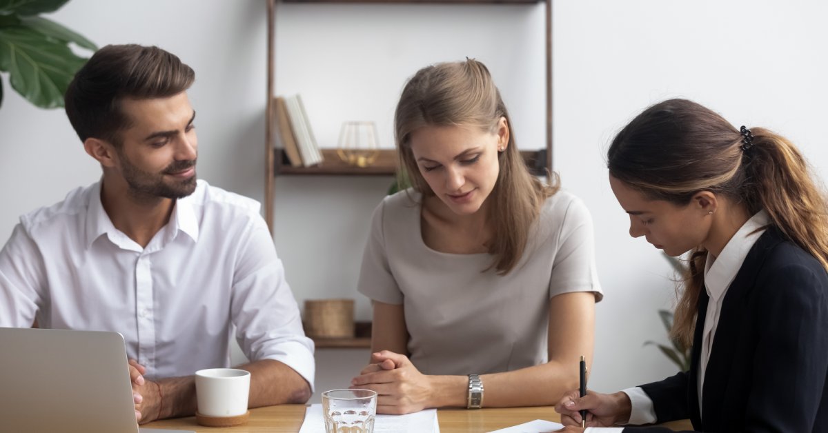 In an office, three people are sitting at a table and are reviewing documents. A laptop and a glass cup sit on the table.
