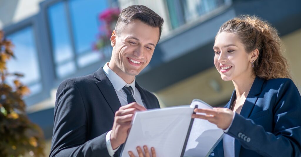 Two business professionals are smiling, standing in front of a building, and looking at a transparent folder full of paper.