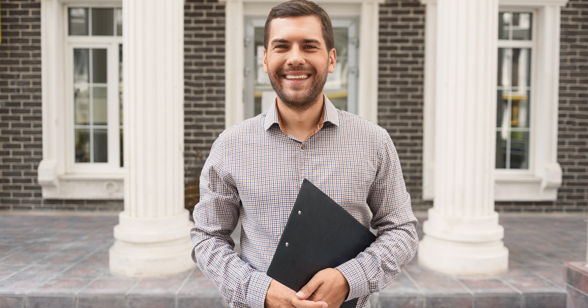 A man wearing a button-down shirt holds a clipboard. He's standing in front of a brick building with white columns.