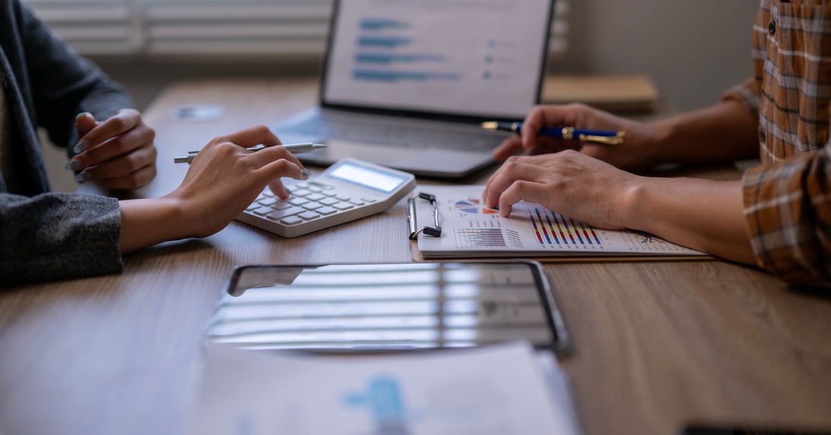 Two people sit across from each other at a desk with a calculator, a clipboard, a tablet, and an open laptop.