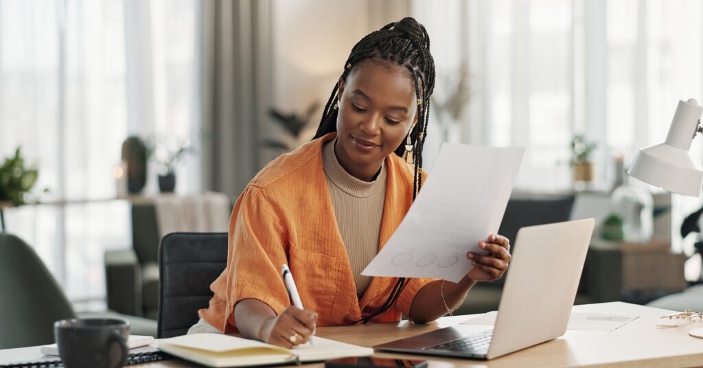 A woman sits at a desk, writes in a notebook, and holds a sheet of paper. A laptop and a mug are on the desk.