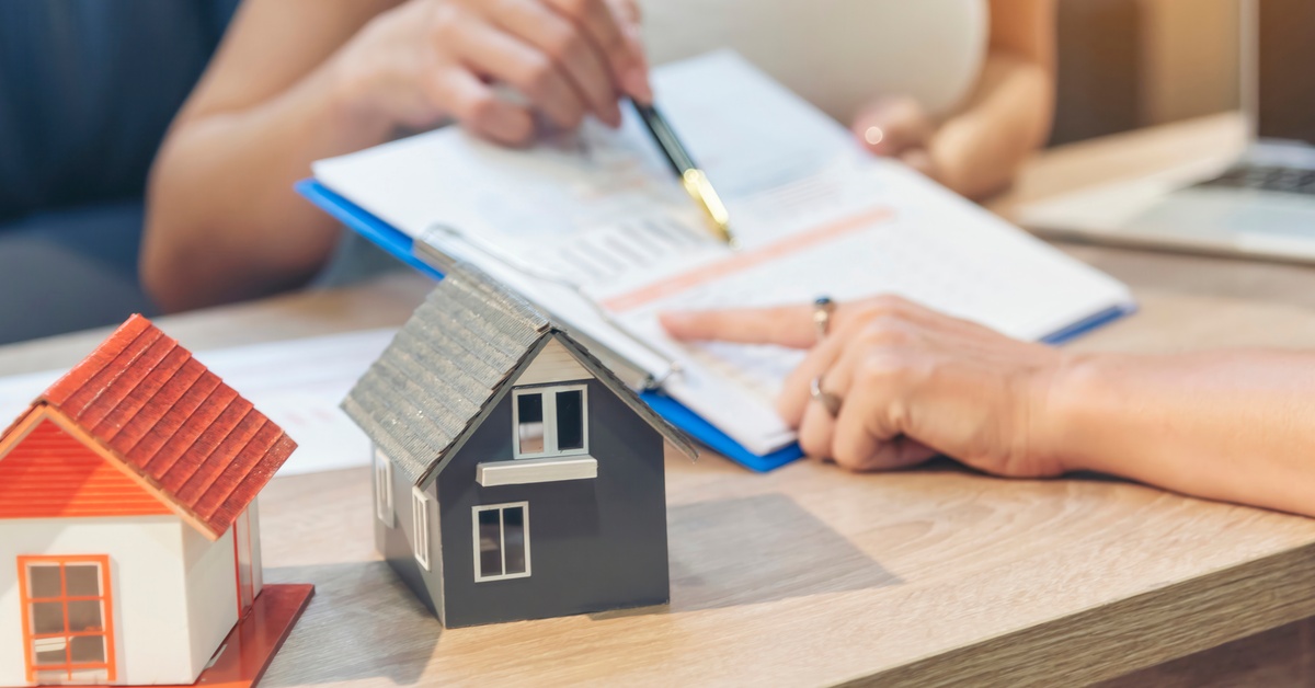 A desk with two house models. One person holds a clipboard and shows highlighted text, and one person points to the text.