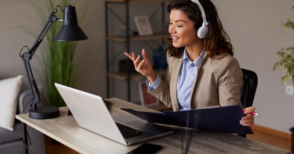 A woman wearing headphones is sitting at a desk with a laptop open. She's holding a folder and speaking.