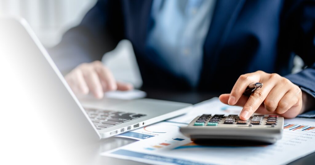 A desk with a laptop, papers that feature graphs, and a calculator. A person presses buttons on the calculator.