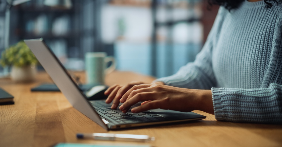 A person wearing a blue sweater sits at a wooden table and types on a laptop. A mug is in the distance.
