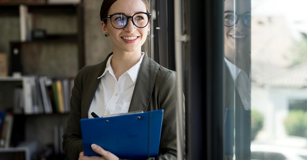 A woman wearing glasses and a blazer holds a blue clipboard while standing. She smiles and looks out the window.